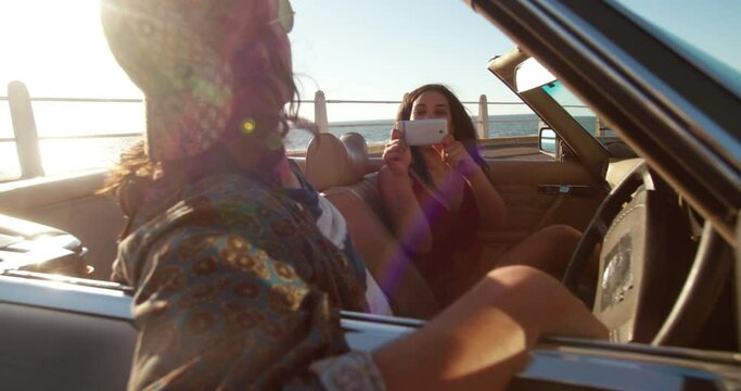 Hipster girl taking pictures to her boyfriend with a smartphone sitting in a convertible parked close to the beach