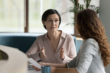 Confident mature business professional woman talking to younger female colleague at office table, speaking, gesturing, teaching, explaining work tasks. Elder mentor training intern - obrazy, fototapety, plakaty