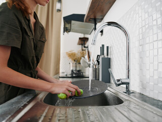 Woman with hands without gloves washes dishes with a dishwashing sponge, household chores, no dishwasher, high water consumption. Close-up of hands