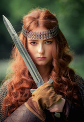 Beautiful red haired girl in metal medieval armor dress with sword standing in warlike pose and...