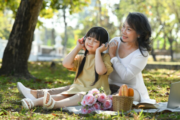 Cute little Asian girl in headphones listening to music while picnic with grandmother at summer park. Family, leisure concept