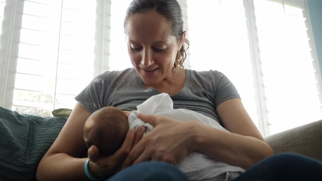 Low angle handheld shot of mother breastfeeding son while sitting on sofa against windows at home