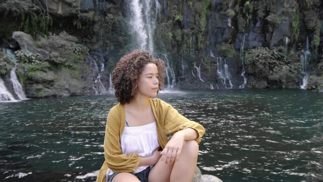Zoom out shot of thoughtful woman looking away while sitting on rock against waterfall