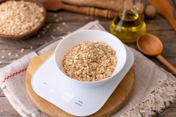 Oatmeal in a white bowl on the kitchen scales on wooden background, rustic style, weight 100 g
