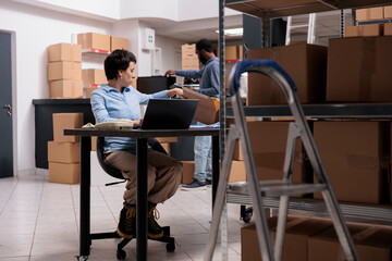 Caucasian worker sitting at desk analyzing customer shipping detalies on laptop computer before start preparing package, working on delivery in warehouse. Distribution center company