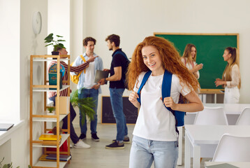 Portrait of a happy school girl with a backpack. Beautiful redhead teenage student girl in a white T shirt and jeans standing in the classroom, with a group of other students in the background
