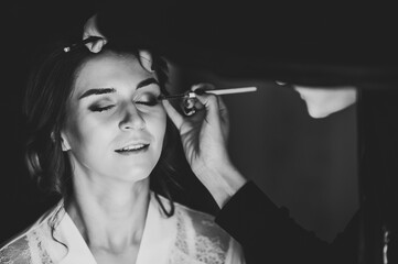 Make-up artist applying eyeshadow on model's eye and holding a brush on background, close up. Young beautiful woman. Black and white photo.