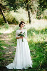 Obraz na płótnie Canvas Young beautiful girl in elegant dress is standing and holding hand bouquet of white flowers and greens with ribbon at nature. The bride holds a wedding bouquet outdoors.