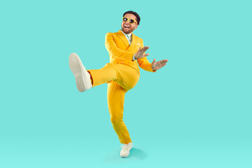Fototapeta na wymiar Full length portrait of happy cheerful young man in yellow suit and sunglasses dancing on turquoise background. He is rising one leg and smiling. Banner for advertisement, marketing with copy space.