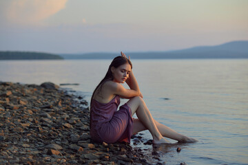 Fototapeta na wymiar Beautiful young woman sitting on shore of lake in summer dress at sunset. Portrait of a romantic wet girl at sunset, warm sun, natural beauty of a woman
