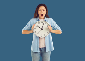 Shocked stunned wondered brunette woman in casual clothes blue shirt, jeans and white t-shirt is holding clock in her hands on blue background. Wasting time, late, empty life, crisis concept.