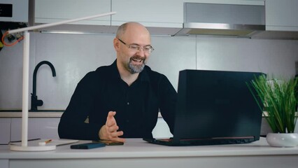 Handsome laughing caucasian man working on laptop computer while sitting behind desk in cozy kitchen in small flat. Busy worker freelancer entrepreneur working home at kitchen in financial sphere - Powered by Adobe