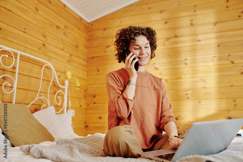 Poster young smiling woman in pajamas speaking on mobile phone and networking in front of laptop while sitt