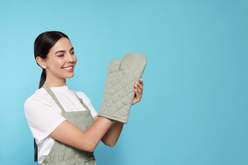 Young woman in grey apron and oven glove on light blue background, space for text