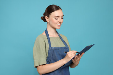 Beautiful young woman in clean denim apron with clipboard on light blue background