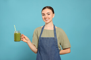 Beautiful young woman in denim apron with delicious smoothie on light blue background