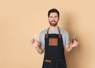 Smiling hairdresser in apron holding scissors on light brown background, space for text