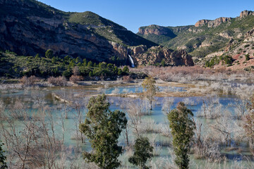 Fototapeta na wymiar Beautiful landscape of the waterfall near the old town of Domeño on a rocky mountain full of vegetation and the Turia river with three trees in the foreground, in Valencia, Spain