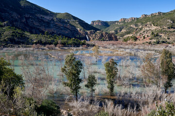 Fototapeta na wymiar Beautiful landscape of the waterfall near the old town of Domeño on a rocky mountain full of vegetation and the Turia river with a lot of leafless groves and three trees in the foreground, in Valencia
