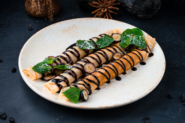 Sweet thin pancakes with cream cheese, banana, chocolate sauce, blueberries, mint and powdered sugar on a plate.