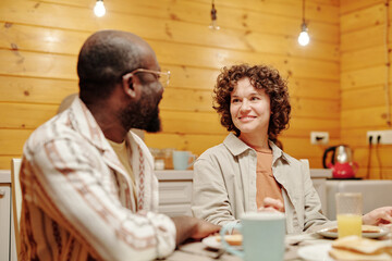 Young smiling brunette woman looking at her African American husband while sitting in front of him by served table during breakfast