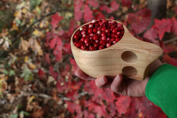 Woman holding tasty lingonberries in wooden cup near red leaves outdoors, closeup - Powered by Adobe