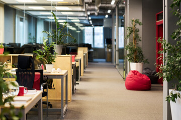 Part of spacious office with long aisle and row of desks with business supplies and green plants standing by walls and on workplaces