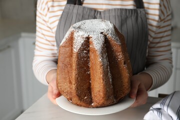 Woman holding delicious Pandoro cake decorated with powdered sugar in kitchen, closeup. Traditional...