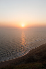 Obraz premium Sunset or sunrise on wild beach with sandy coastline and grassy hills, red sun in misty sky leaving light on water surface. Tranquility and beauty of nature. Perfect place for meditation