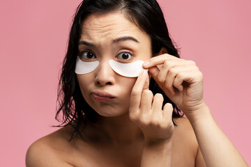 Cute beautiful woman takes care of skin, has under eye patches over pink background