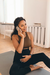 Fototapeta na wymiar Vertical image of pregnant african american woman sitting on mat in sportswear talking on phone with her doctor, asking if she can practice prenatal yoga and doing special asanas, stroking her belly