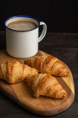 Top view of croissants on board on wooden table with rustic cup of coffee, black background, vertical, with copy space