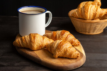 Top view of croissants on board on wooden table with rustic cup of coffee, black background, horizontal, with copy space