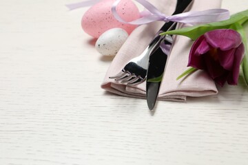 Cutlery set, painted eggs and beautiful flower on white wooden table, space for text. Easter celebration