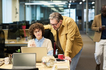 Young confident businesswoman in casualwear explaining something to mature male colleague while both looking at laptop screen