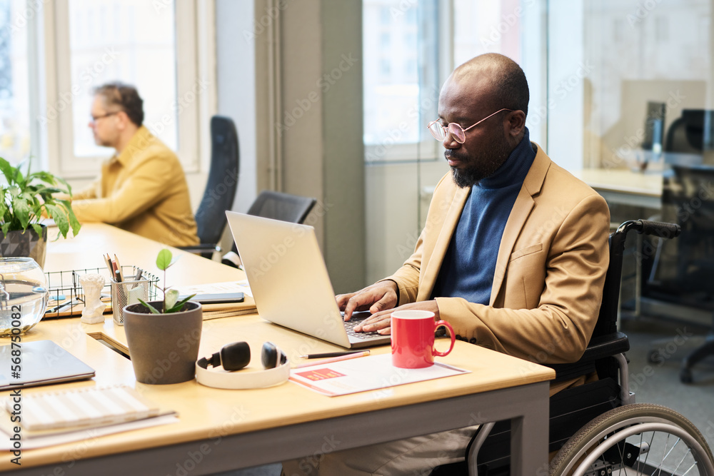 Poster serious young male broker with disability typing on laptop keypad while sitting by workplace in cowo