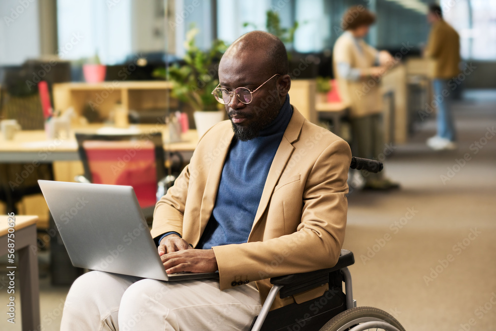 Poster serious young businessman with laptop sitting in wheelchair in openspace office and looking attentiv