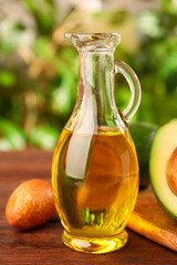 Fresh avocados and jug of cooking oil on wooden table against blurred green background, closeup