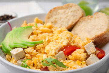 Bowl with delicious scrambled eggs, tofu and slices of baguette, closeup