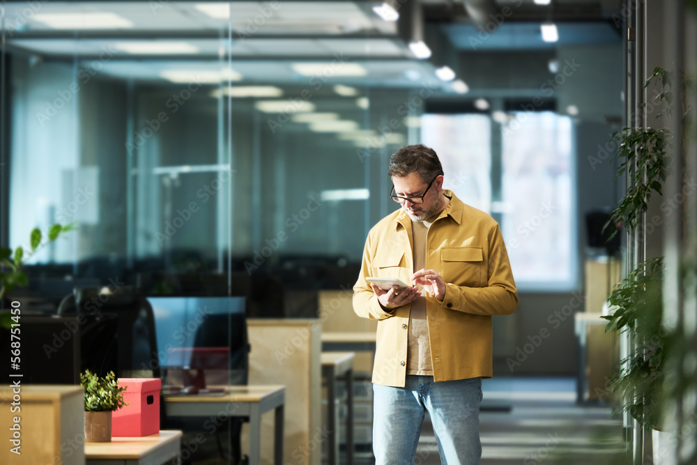 Poster mature male broker in casual attire using tablet while standing in aisle in openspace office and loo