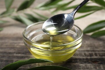 Pouring olive oil from spoon into glass bowl on wooden table, closeup