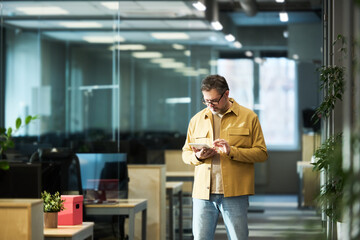 Mature male broker in casual attire using tablet while standing in aisle in openspace office and...