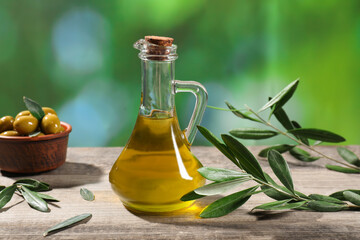 Jug of cooking oil, olives and green leaves on wooden table against blurred background