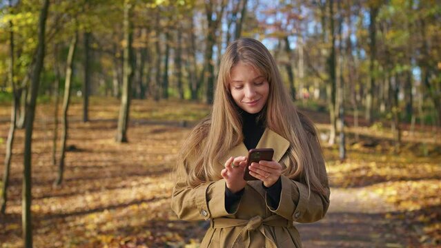 Front view of happy, cheerful woman enjoying sunny autumn weather in park. Attractive, young lady standing, using smartphone, closing eyes, smiling. Concept of beauty of nature.