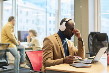 Young tired businessman in headphones sitting by workplace in front of laptop and having short break against his colleagues having talk