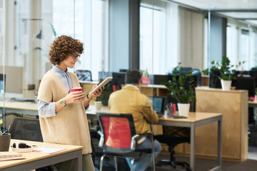 Obraz premium Young female manager with cup of coffee and smartphone texting or scrolling through online posts against male colleague with laptop