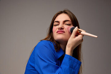 Obraz premium Half-length portrait of young emotional beautiful girl holding blush brush and doing makeup isolated over grey background. Beauty, fashion, style, fun and joy concept