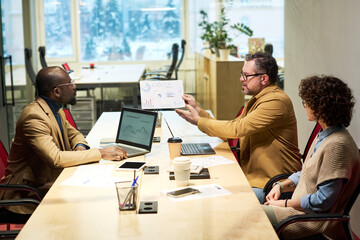 Confident mature male analyst showing paper document with graphic financial data to young...