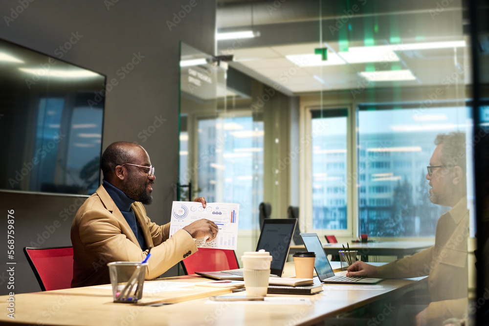 Poster confident male economist commenting graphic data on paper document while sitting by desk in front of