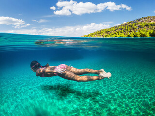 Girl diver is snorkeling on a beautiful sea beach. The lower half of the image is occupied by the seabed, the upper half by the coast and the sky.